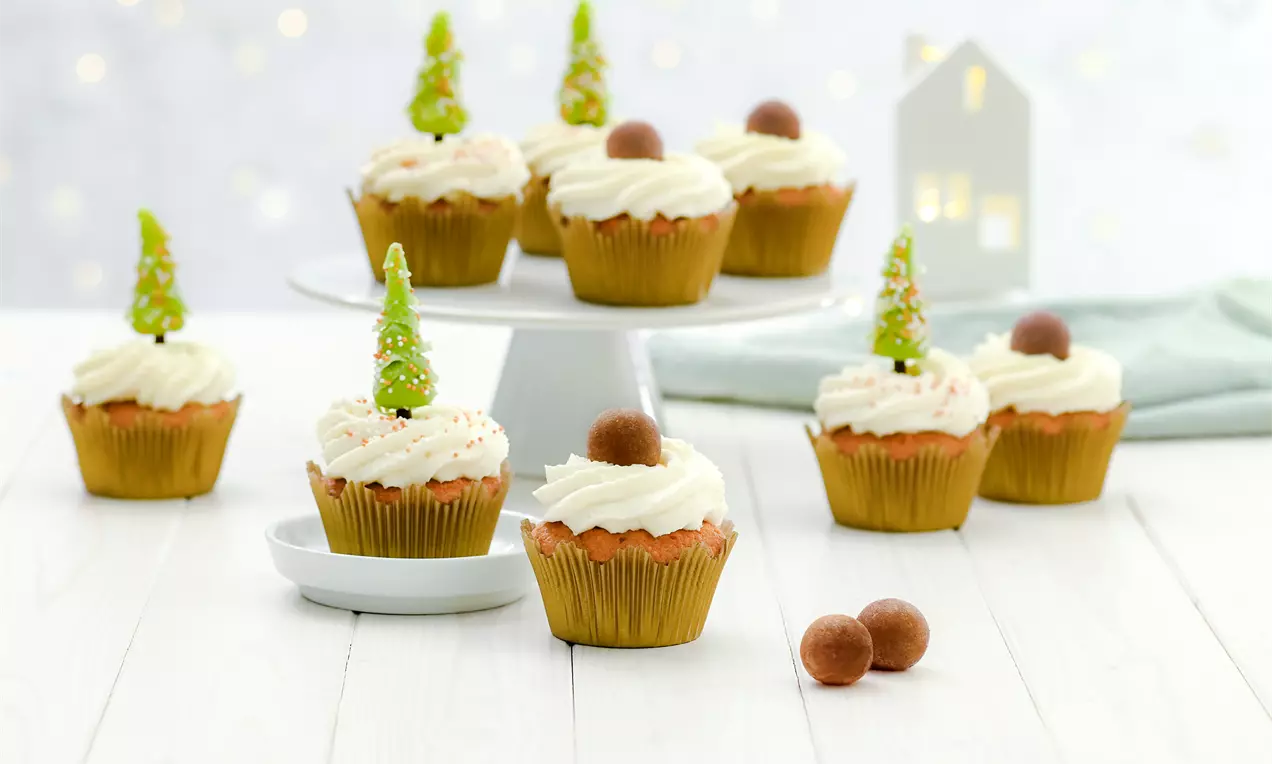 Aranybarna, puha muffin hófehér krémkoronával, marcipángolyóval és zöld fenyődíszekkel, fehér tányérokon világos háttér előtt