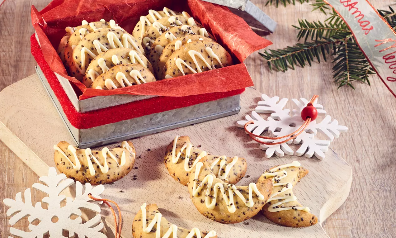 Aranyszínű, ívelt kiflik vaníliával és chia magokkal, fehér cukros mázzal díszítve, fenyőág és hópehely dekoráció mellett fa tálcán