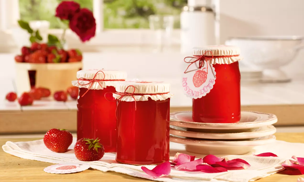Három, fényesen piros eperzselével töltött befőttesüveg fehér díszpapírral és madzaggal átkötve, friss eperrel és rózsaszirmokkal körítve.