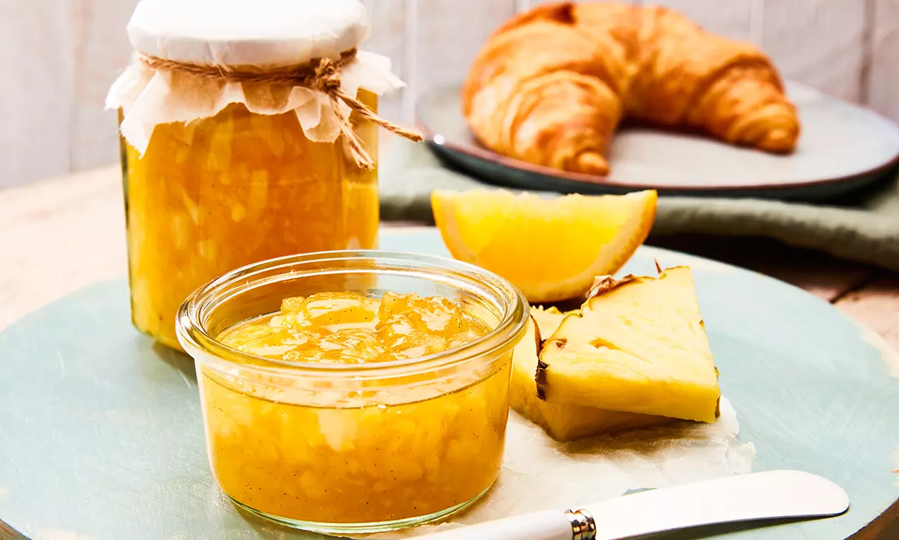 Világos, aranyló színű ananász-narancsdzsem üvegedényben, mellette friss ananászszeletek és narancsgerezd egy kék tányéron croissanttal