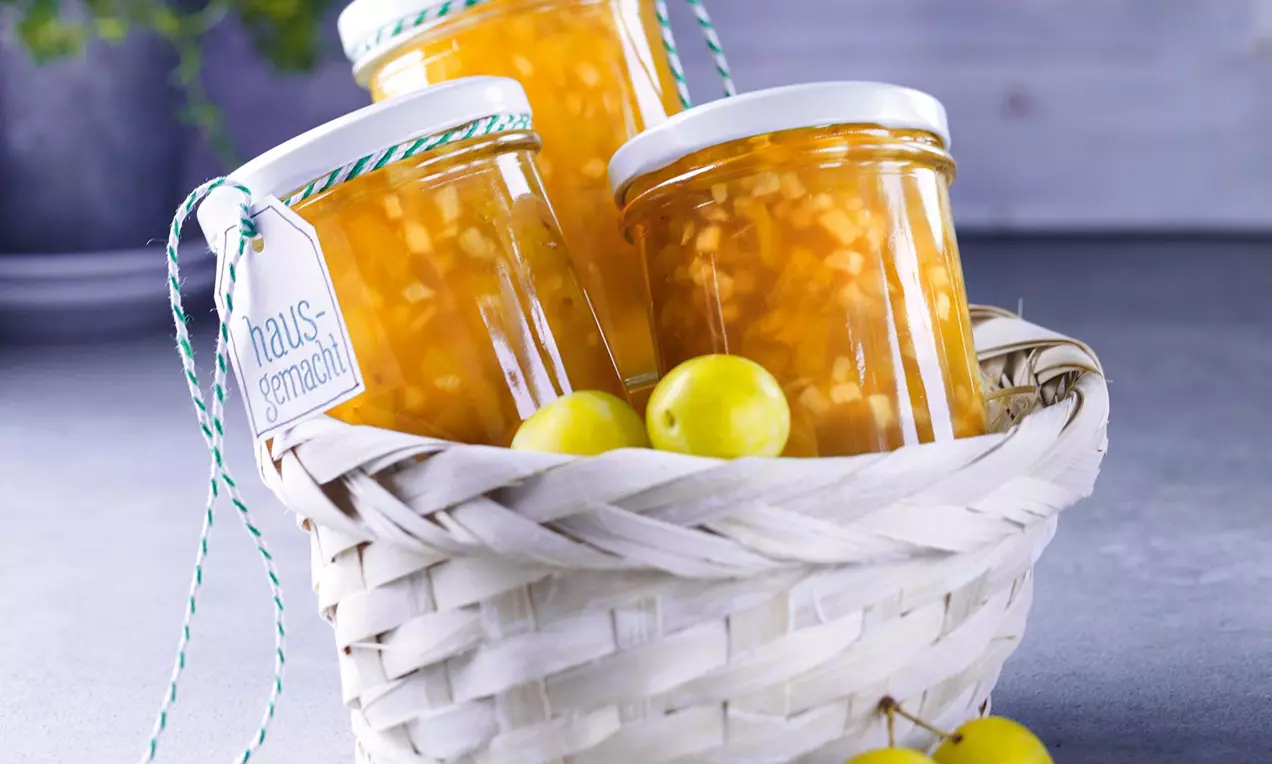 Három aranyló szilva-darabokkal teli lekváros üveg fehér fedéllel és zöld-fehér zsinórral fonott kosárban sárga mirabella szilvák között