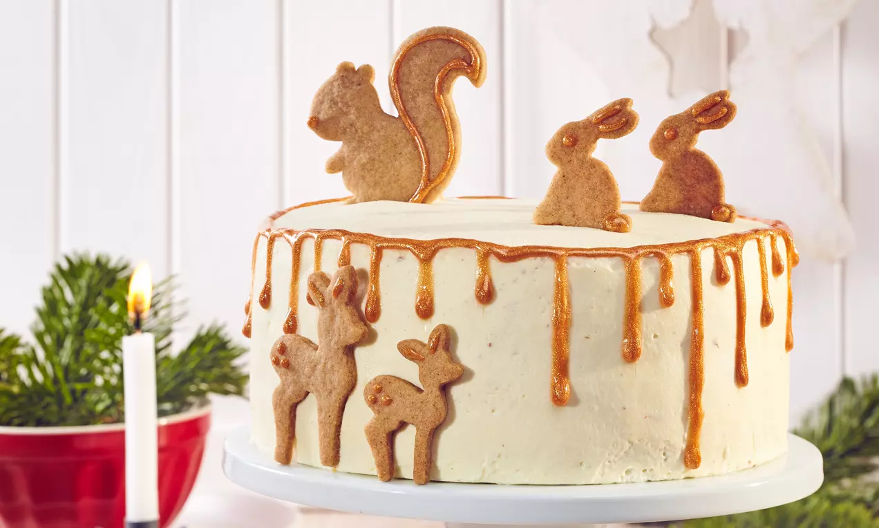 Fehér, enyhén rusztikus torta karamellcsíkokkal, tetején aranybarna mézeskalács figurák, körülötte ünnepi gyertyák és fenyőágak