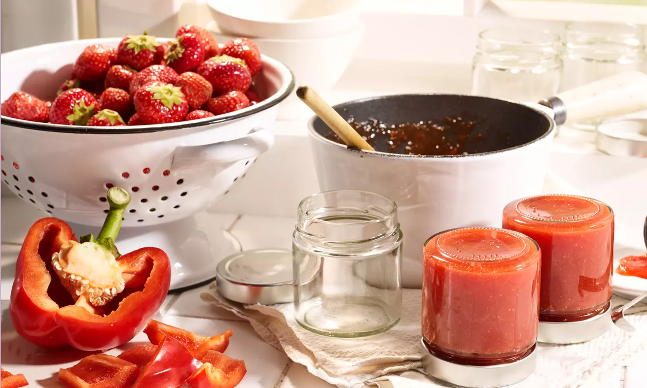 Élénkpiros eper és paprika mellett üvegek, sűrű piros krémmel töltve, fehér szűrő, és fazék rendezett konyhai háttérben